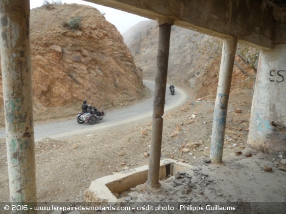 Des motos passent sur la route 203 devant une ruine